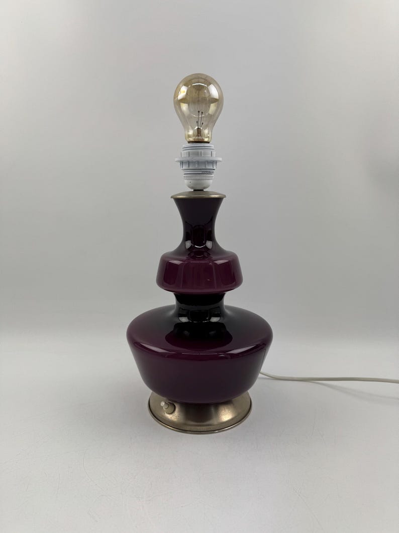 May include: A purple glass table lamp with a silver base and a light bulb. The lamp has a layered design with a wide base and a narrow neck.