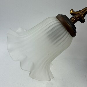 May include: A white, ruffled glass lampshade with a brass-colored metal base and decorative finial. The shade has a bell-like shape with vertical ridges and a scalloped edge. The finial has an ornate, floral design.