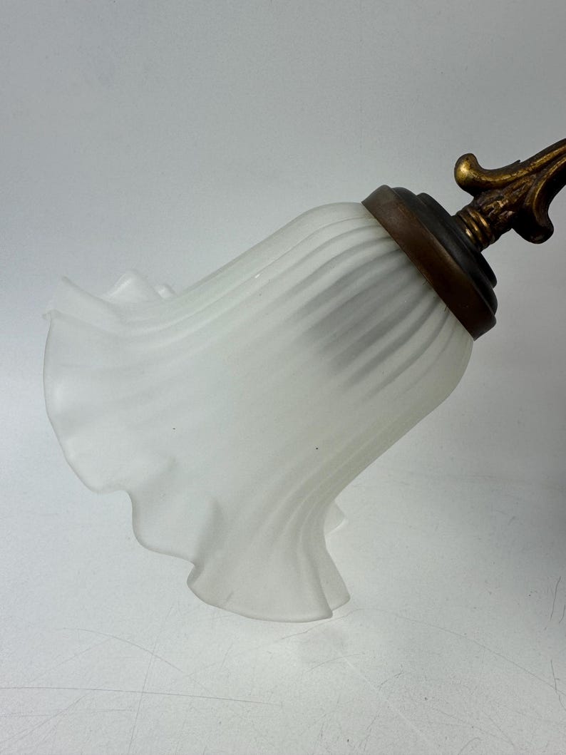May include: A white, ruffled glass lampshade with a brass-colored metal base and decorative finial. The shade has a bell-like shape with vertical ridges and a scalloped edge. The finial has an ornate, floral design.