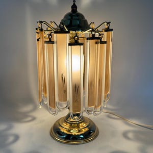 Op de afbeelding: Een vintage tafellamp met een goudkleurige voet en accenten. De lamp heeft een donkergroene bovenkant en een cilindrische kap bestaande uit verticale, rechthoekige panelen. De panelen zijn licht amberkleurig met een bladmotief. De lamp is verlicht en werpt schaduwen.