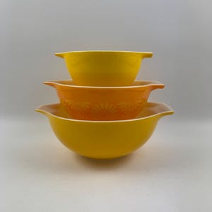 Op de afbeelding: Een set van drie gele mengkommen, de kleinste kom is geel met een witte rand, de middelste kom is oranje met een witte rand en een bloemenpatroon, en de grootste kom is geel met een witte rand.