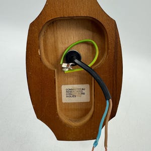 May include: A wooden wall sconce back with wiring and a label. The sconce is a light brown color and has a curved shape. The wiring includes a black cable, a green wire, a blue wire, and a brown wire.