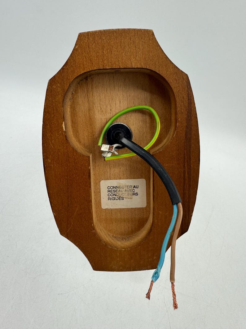 May include: A wooden wall sconce back with wiring and a label. The sconce is a light brown color and has a curved shape. The wiring includes a black cable, a green wire, a blue wire, and a brown wire.