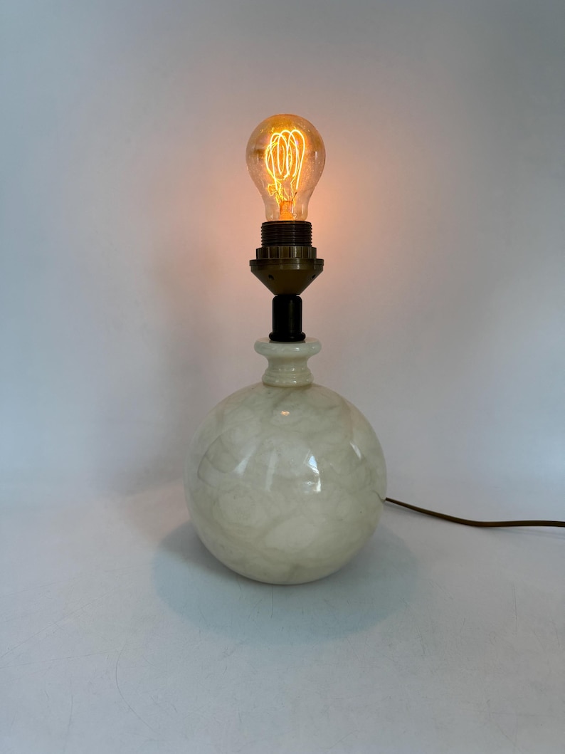 Peut inclure: Une lampe de table vintage avec une base sph&eacute;rique &agrave; effet marbr&eacute;. La base est blanc cr&egrave;me avec un motif tourbillonnant. L'ampoule d&eacute;corative est allum&eacute;e, d&eacute;gageant une lumi&egrave;re orange chaude. Un c&acirc;ble noir part de la base.