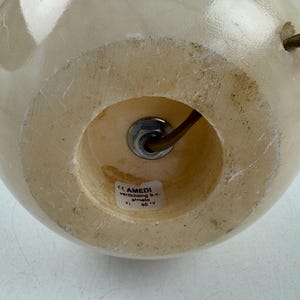 Peut inclure: Base de lampe sph&eacute;rique de couleur cr&egrave;me, avec une ouverture circulaire en bas. Un &eacute;crou m&eacute;tallique fixe un fil marron &agrave; l'int&eacute;rieur. La base a une surface lisse et polie. Le texte "AMEDI verlichting b.v. almelo" est visible sur une &eacute;tiquette.
