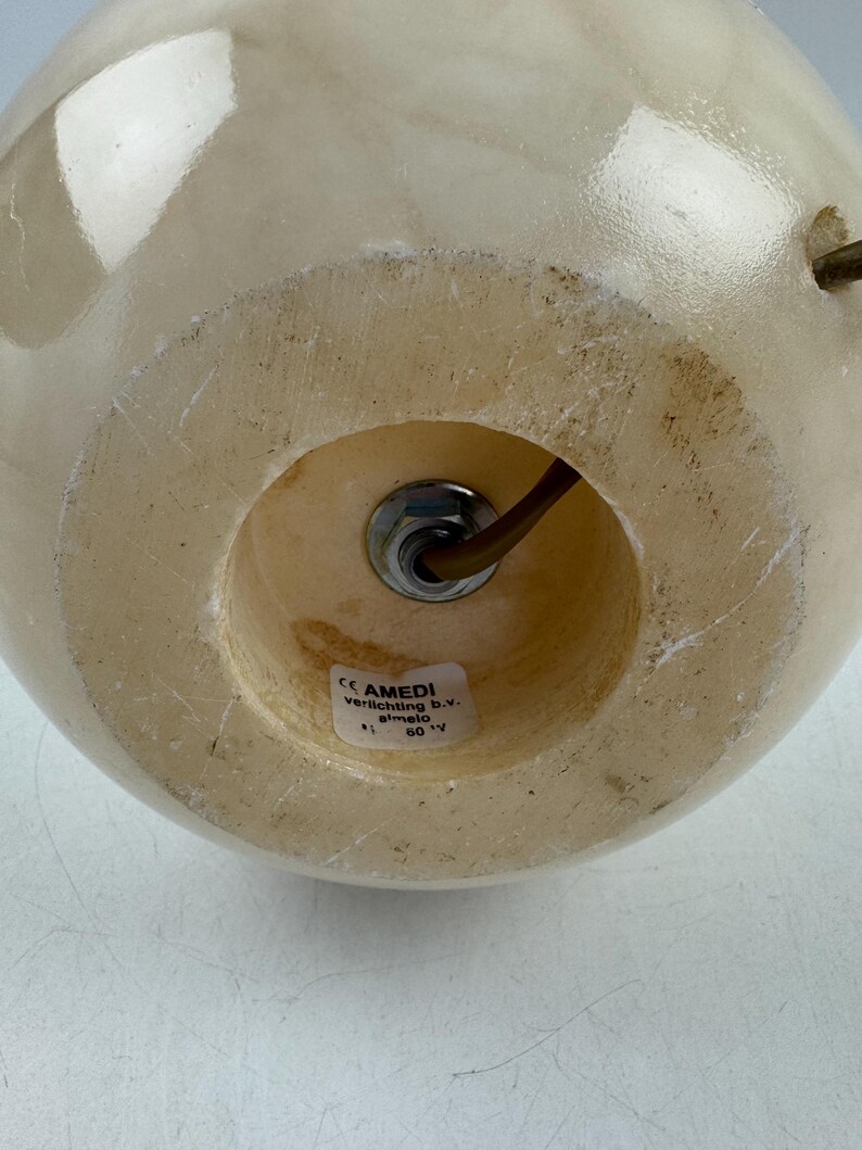 Peut inclure: Base de lampe sph&eacute;rique de couleur cr&egrave;me, avec une ouverture circulaire en bas. Un &eacute;crou m&eacute;tallique fixe un fil marron &agrave; l'int&eacute;rieur. La base a une surface lisse et polie. Le texte "AMEDI verlichting b.v. almelo" est visible sur une &eacute;tiquette.