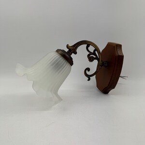 May include: A vintage wall sconce featuring a white, ruffled glass shade and a decorative brass arm. The sconce is mounted on a dark brown wooden base. The fixture has a classic, elegant design.