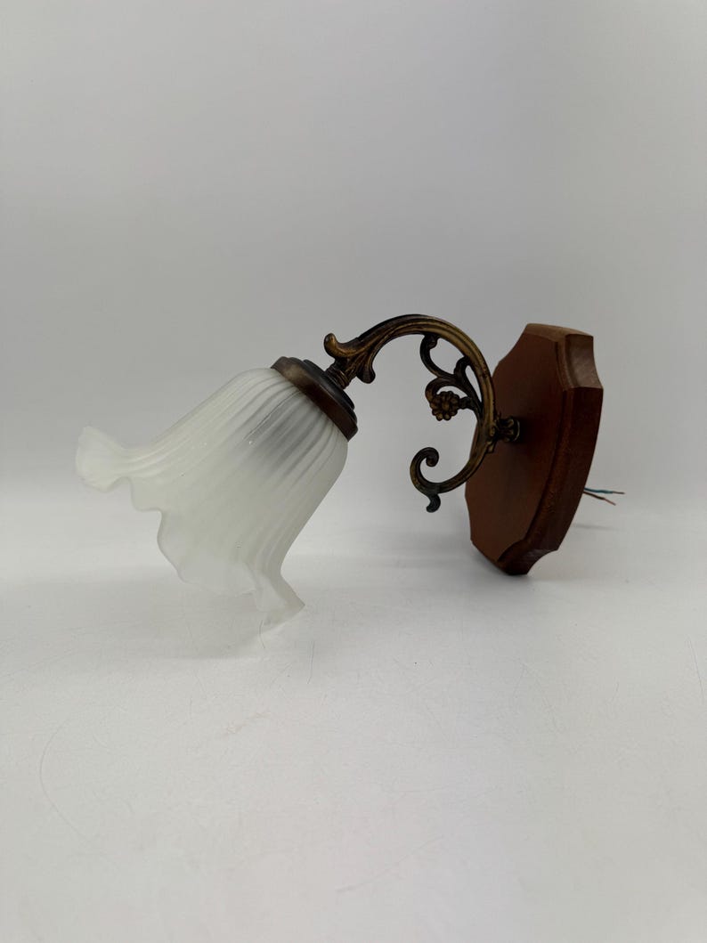 May include: A vintage wall sconce featuring a white, ruffled glass shade and a decorative brass arm. The sconce is mounted on a dark brown wooden base. The fixture has a classic, elegant design.