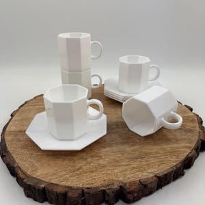 May include: A set of white, geometric-shaped teacups and saucers. The cups are stacked and arranged on a wooden serving board. The cups have a handle and a faceted design. The saucers are square with a geometric shape. The background is white.