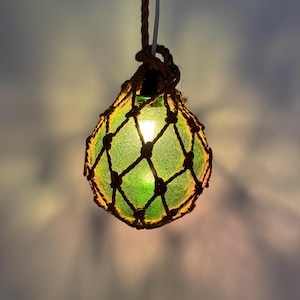 Op de afbeelding: Een groene glazen bol omsloten door een natuurlijk touwnet, hangend aan een wit snoer. De bol wordt van binnenuit verlicht, waardoor een warme gloed ontstaat.