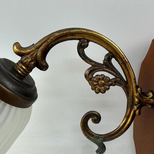 May include: An antique wall sconce with a decorative brass arm featuring floral and scrollwork details. The sconce has a white, ribbed glass shade and a dark brown metal base. The brass has a gold finish with some areas of green oxidation.