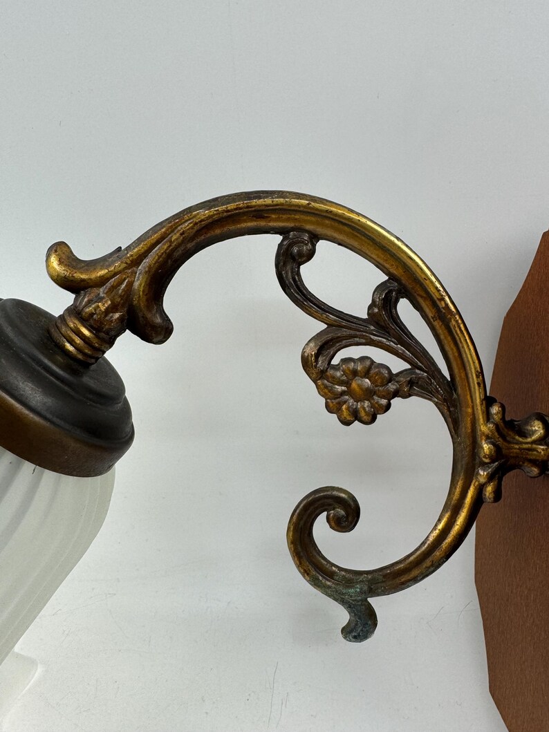 May include: An antique wall sconce with a decorative brass arm featuring floral and scrollwork details. The sconce has a white, ribbed glass shade and a dark brown metal base. The brass has a gold finish with some areas of green oxidation.