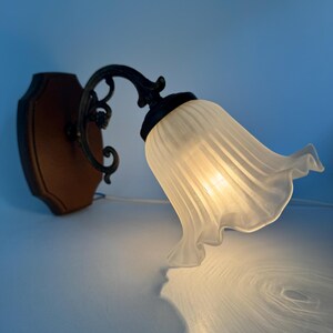 May include: A vintage wall sconce with a brown wooden base and a decorative black metal arm. The sconce features a frosted glass shade shaped like a flower, casting a soft light. The design is reminiscent of antique lighting fixtures.