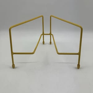 Op de afbeelding: Een gele metalen plantenstandaard met een geometrisch ontwerp. De standaard heeft een rechthoekige basis en schuine steunen, waardoor een open structuur ontstaat. De standaard is ongeveer 30 cm breed en 25 cm hoog.