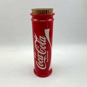 Op de afbeelding: Een rode cilindrische Coca-Cola-container met een kurken deksel. De container heeft het Coca-Cola-logo en de woorden "Enjoy" en "Reg. Trademark" erop gedrukt.