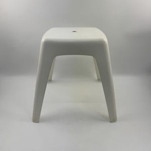 Puede incluir: Un taburete de plástico blanco con un asiento cuadrado y cuatro patas anguladas. El taburete tiene un diseño simple y minimalista y es adecuado para uso en interiores o exteriores. El taburete mide aproximadamente 43 cm de alto.