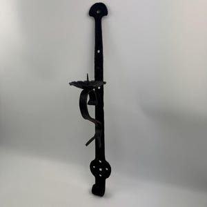Pode incluir: Arandela de parede em ferro forjado preto com um design decorativo. A arandela apresenta um topo circular com um orifício para pendurar, um castiçal e uma base decorativa com um design curvo. Projetado para uma única vela.