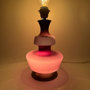 May include: A pink and purple glass table lamp with a white frosted shade and a round base. The lamp is lit with a warm white light bulb.