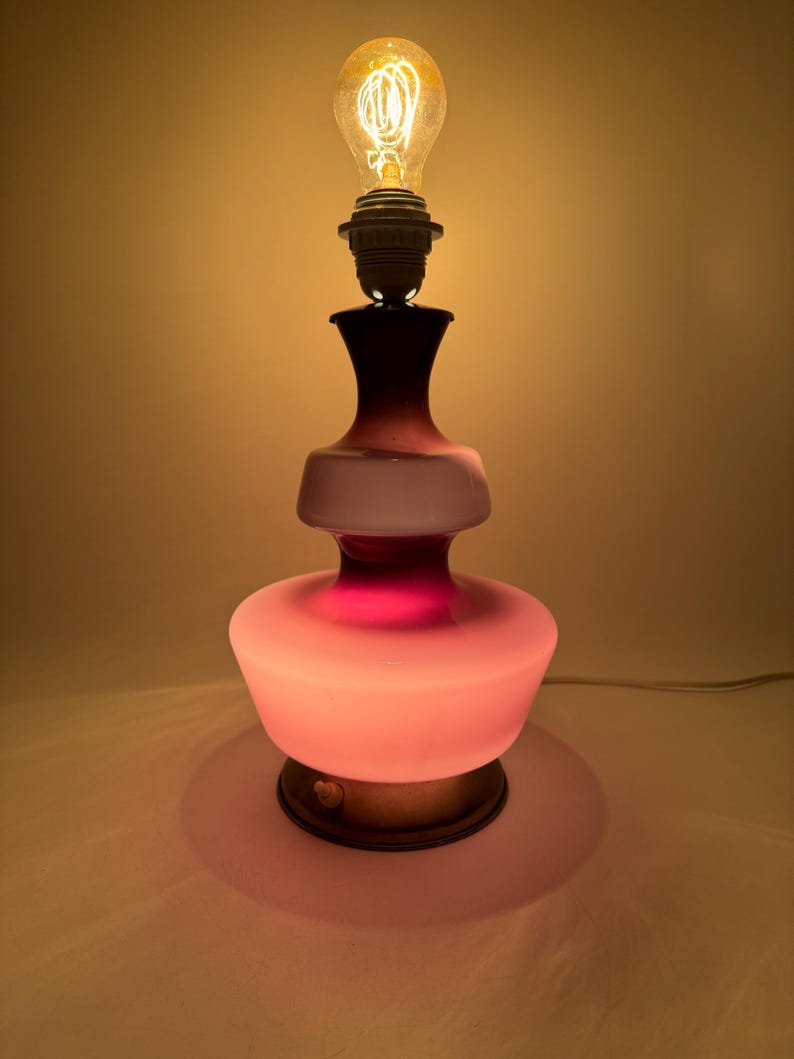 May include: A pink and purple glass table lamp with a white frosted shade and a round base. The lamp is lit with a warm white light bulb.