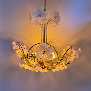Op de afbeelding: Een goudkleurige kroonluchter met witte bloemvormige tinten. De armatuur heeft meerdere armen met witte bloemaccenten en een centrale cluster van decoratieve elementen. Het ontwerp is sierlijk en elegant, geschikt voor binnenverlichting.