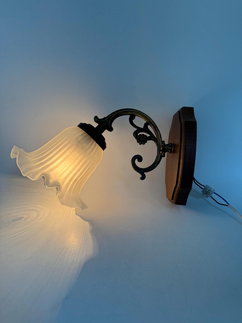 May include: A wall-mounted light fixture with a frosted glass shade shaped like a flower. The fixture has a dark metal arm with decorative flourishes and a wooden base. The light is on, casting a warm glow.