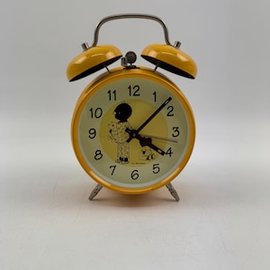 Könnte beinhalten: Gelber Wecker mit einer schwarzen Silhouette einer Person im Schlafanzug, die einen Teddybär hält. Das Zifferblatt der Uhr hat schwarze Zahlen und Zeiger. Die Uhr hat eine silberne Glocke und einen silbernen Griff.
