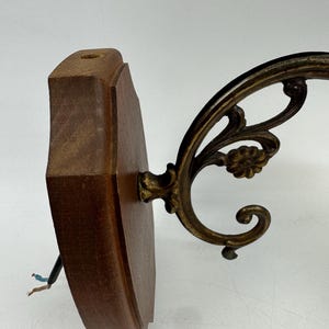 May include: A vintage wall sconce with a dark wood backing and ornate brass-colored metal arm. The sconce features a decorative floral design and exposed wiring. The wooden base is approximately 6 inches tall.