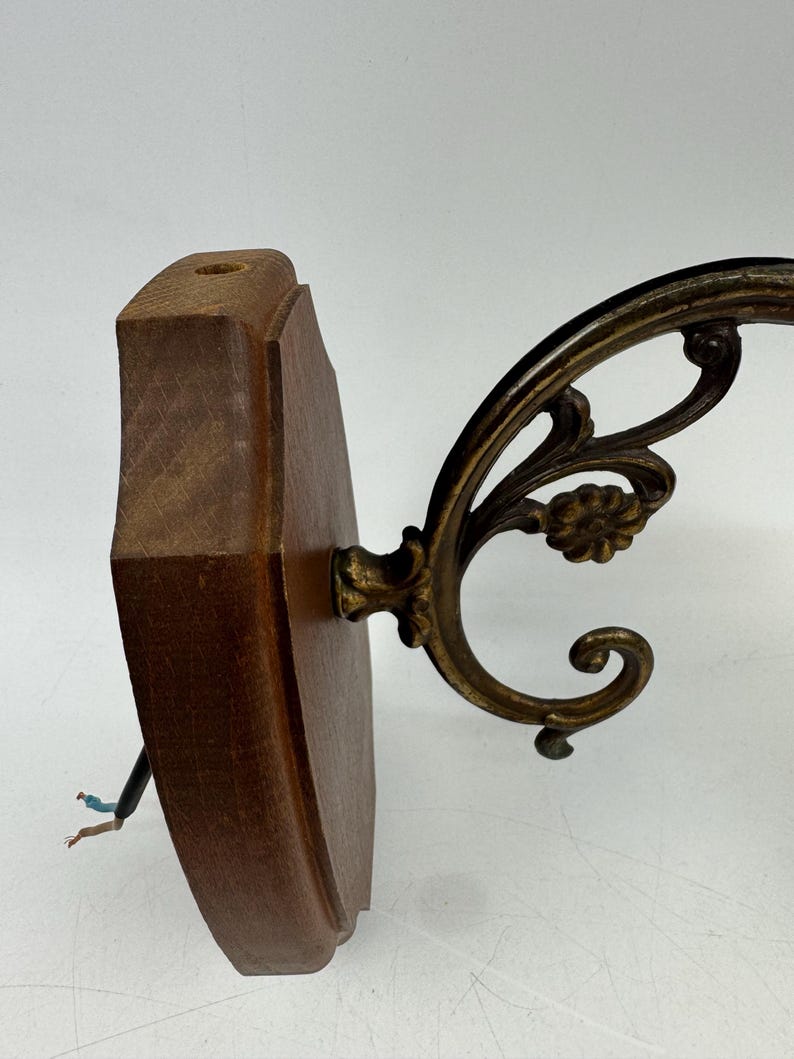 May include: A vintage wall sconce with a dark wood backing and ornate brass-colored metal arm. The sconce features a decorative floral design and exposed wiring. The wooden base is approximately 6 inches tall.