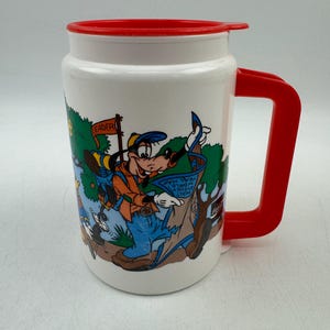 Op de afbeelding: Witte reismok met een rode deksel en handvat, met een cartoonafbeelding van Goofy in een bosscène. De mok heeft de tekst "Leader" op een vlag en "Walt Disney World Trail Map".