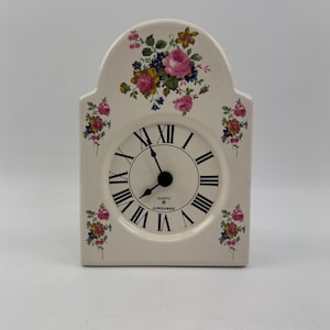 Op de afbeelding: Crèmekleurige quartz klok met een bloemenontwerp. De wijzerplaat heeft zwarte Romeinse cijfers en zwarte wijzers. De klok is rechthoekig met een gewelfde bovenkant en bloemaccenten.