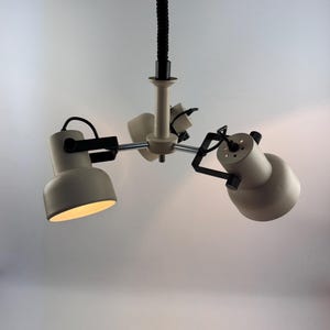 Op de afbeelding: Een moderne plafondlamp met drie lampen in een crèmekleurige afwerking. De lamp heeft drie verstelbare spots met zwarte accenten en een flexibel zwart snoer. De lampen zijn naar beneden gericht en verlichten de ruimte.