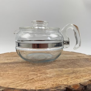 Könnte beinhalten: Eine klare Glasteekanne mit rundem Körper, gebogenem Griff und Deckel. Ein silbernes Band umschließt die Mitte der Teekanne. Die Teekanne steht auf einer runden Holzoberfläche mit natürlichem Rand.
