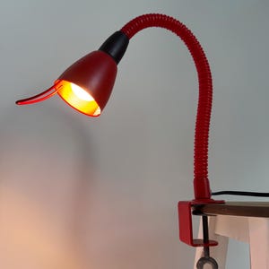 Puede incluir: Lámpara de escritorio roja con diseño de cuello de cisne flexible. La lámpara tiene una pantalla roja y una junta negra, y está sujeta a una superficie de madera. La lámpara está encendida, emitiendo una luz cálida y amarilla.