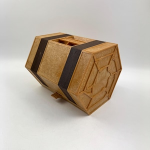 Puede incluir: Una caja hexagonal de madera con acabado marrón claro y bandas marrones oscuras. La caja tiene un diseño geométrico decorativo en los lados y la parte superior. La caja probablemente se utiliza para almacenamiento o exhibición.