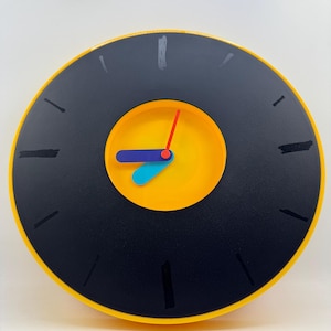 May include: A circular clock with a black face and a yellow border. The clock has black dashes for hour markers and a yellow centre. The hands are red, blue, and yellow. A bright and bold decorative item, suitable for a modern interior.