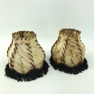 Op de afbeelding: Twee off-white lampenkappen met bruine stiksels en een donkerbruine franje. De kappen hebben een textuur en een uniek, rustiek ontwerp. De kappen zijn waarschijnlijk voor een tafellamp of een soortgelijke armatuur.