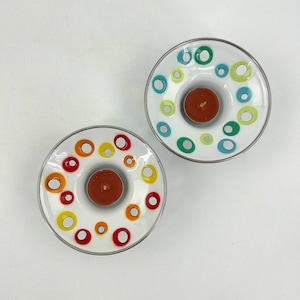Op de afbeelding: Twee witte keramische kaarsenhouders met een zilveren rand. Elke houder heeft een cirkelvormig ontwerp met gekleurde cirkels in rood, geel, groen en blauw.