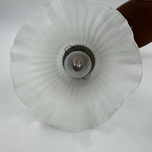 May include: A vintage wall-mounted light fixture with a frosted glass shade. The shade has a ruffled edge and a ribbed design. The fixture includes a brown wooden backing and a visible lightbulb.