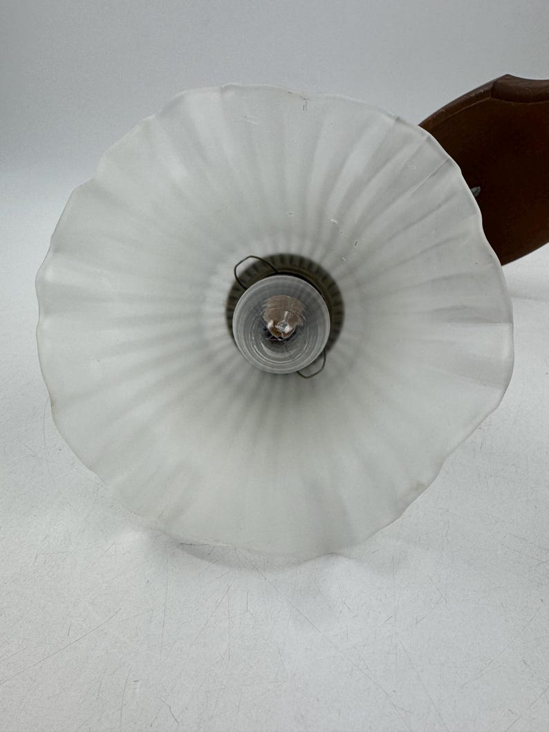 May include: A vintage wall-mounted light fixture with a frosted glass shade. The shade has a ruffled edge and a ribbed design. The fixture includes a brown wooden backing and a visible lightbulb.