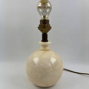 Peut inclure: Une lampe de table vintage avec une base ronde de couleur cr&egrave;me et un col en laiton. La lampe est surmont&eacute;e d'une ampoule transparente. Un cordon &eacute;lectrique marron part de la base. La lampe est un luminaire d&eacute;coratif.