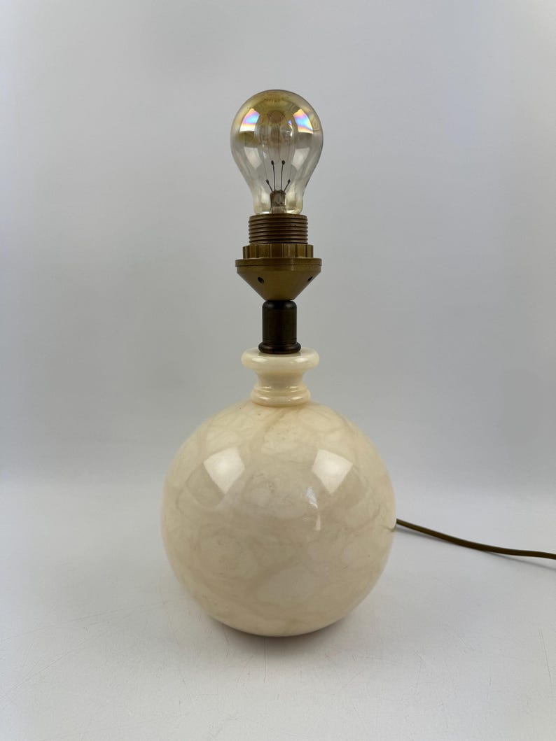 Peut inclure: Une lampe de table vintage avec une base ronde de couleur cr&egrave;me et un col en laiton. La lampe est surmont&eacute;e d'une ampoule transparente. Un cordon &eacute;lectrique marron part de la base. La lampe est un luminaire d&eacute;coratif.