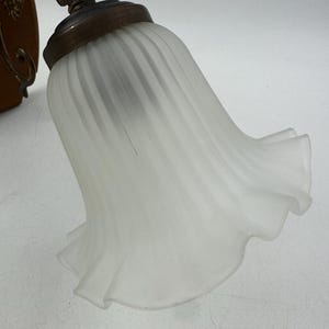 May include: A close-up of a vintage wall sconce with a frosted glass shade. The shade has a ruffled edge and a ribbed design. The metal base has an antique brass finish and decorative details.