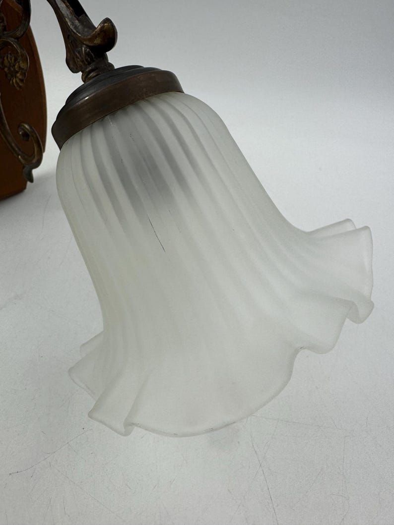 May include: A close-up of a vintage wall sconce with a frosted glass shade. The shade has a ruffled edge and a ribbed design. The metal base has an antique brass finish and decorative details.