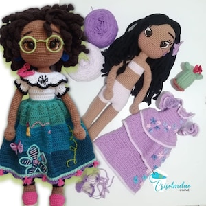 May include: Two crocheted dolls with brown skin and black hair. One doll is wearing a green and white dress with a butterfly design. The other doll is wearing a purple dress with a flower design. Both dolls are holding yarn balls.