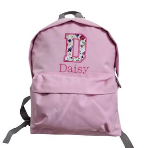 May include: A light pink backpack featuring a large embroidered letter D with a floral pattern in shades of pink, purple, and green. Below the letter, the name Daisy is embroidered in pink thread. The bag has gray straps and a front pocket.