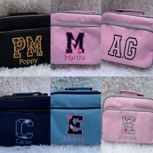 May include: Six personalized lunch bags with different colors and designs. The bags are black, pink, blue, and light blue. Each bag has a large letter embroidered on the front with a name underneath. The letters are "P", "M", "A", "C", "E", and "E". The names are "Poppy", "Martha", "AG", "Carter", "Eleanor", and "Emma".