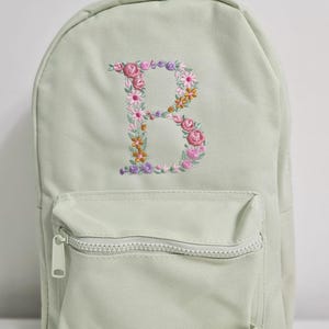 May include: A light green canvas backpack with a zippered front pocket. The backpack has a floral embroidered letter "B" on the front.