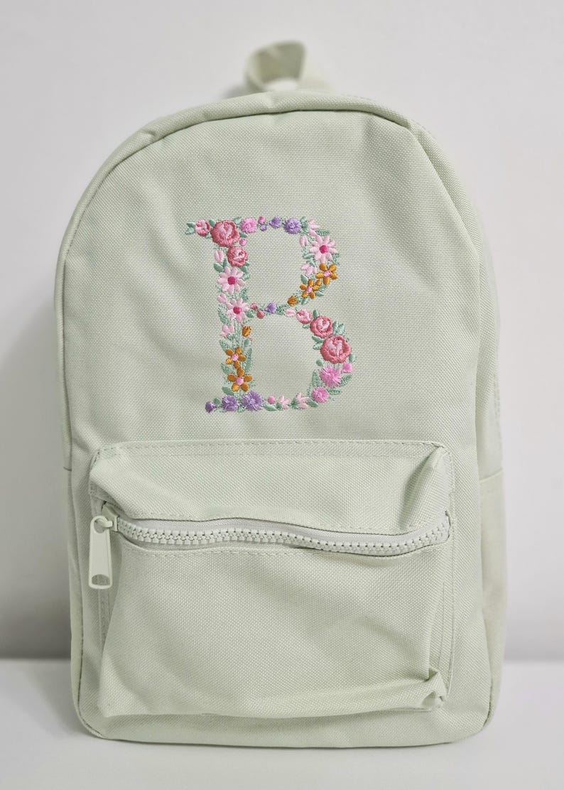 May include: A light green canvas backpack with a zippered front pocket. The backpack has a floral embroidered letter "B" on the front.