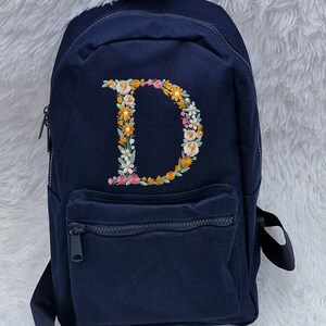 May include: A navy blue canvas backpack with a floral embroidered letter "D" on the front. The backpack has a zippered front pocket and adjustable straps.