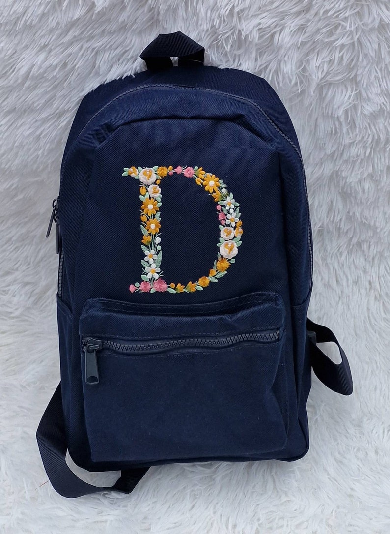 May include: A navy blue canvas backpack with a floral embroidered letter "D" on the front. The backpack has a zippered front pocket and adjustable straps.
